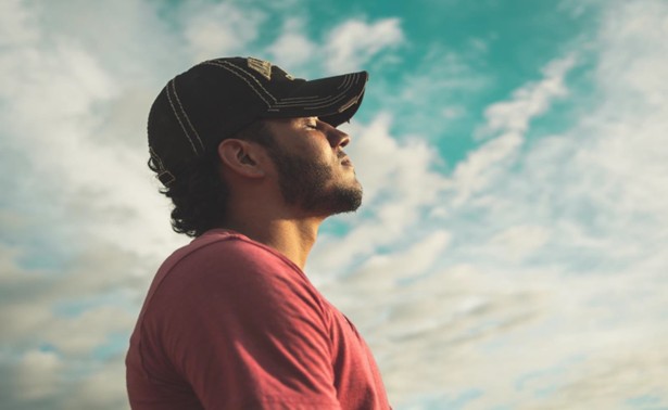 Man standing outdoors with eyes closed and face lifted toward the sky, symbolizing presence, breathwork, calmness, and inner reflection